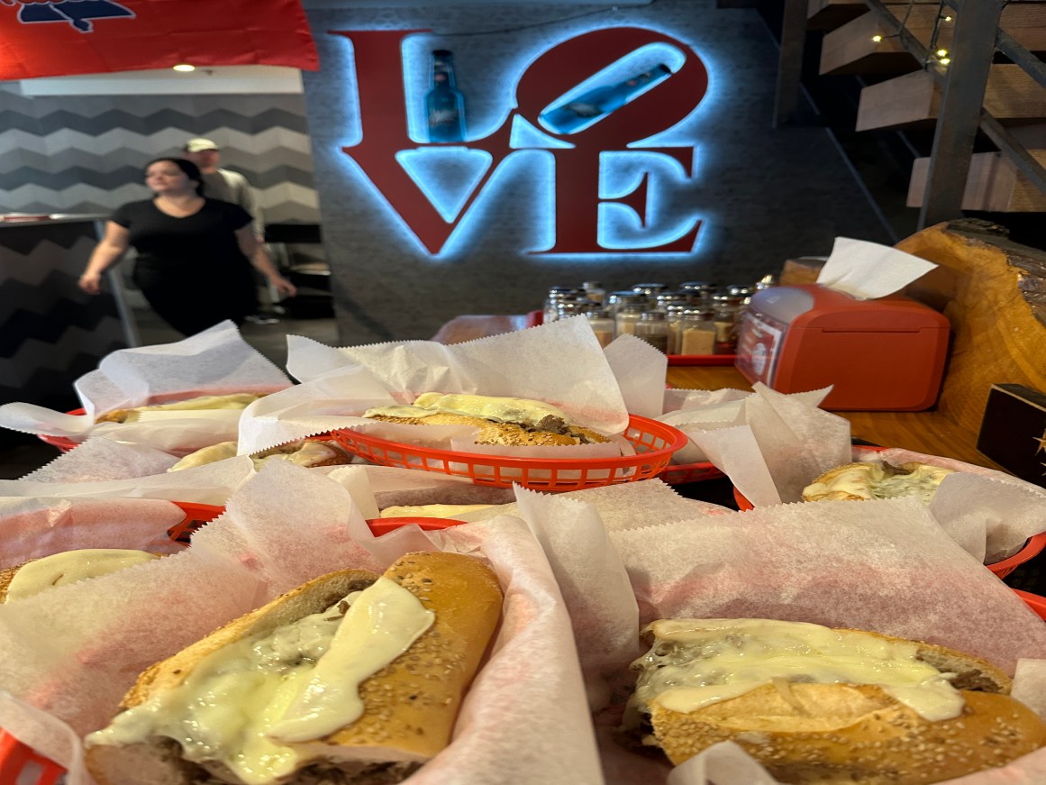 image of A row of Philly cheesesteak sandwiches in red baskets sits on a restaurant table in front of a glowing, blue and red "LOVE" sign